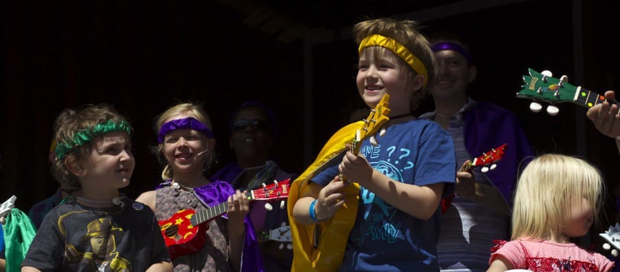 A group of children wearing colourful outfits and accessories play small musical instruments on stage, showcasing a joyful performance interactive installations sounds music