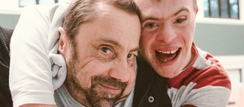 Two dancers from Stopgap Dance Company embrace while looking in to the camera, both have short brown hair, one has a dark beard and is a wheelchair user, they are in an indoor rehearsal room