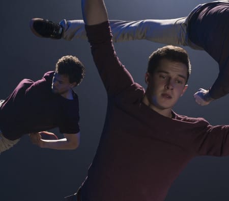 Three acrobats perform dynamic moves within a large hoop, illuminated by dramatic stage lighting against a smoky backdrop.