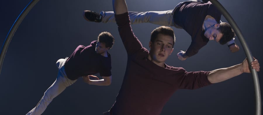 Three acrobats perform dynamic moves within a large hoop, illuminated by dramatic stage lighting against a smoky backdrop.