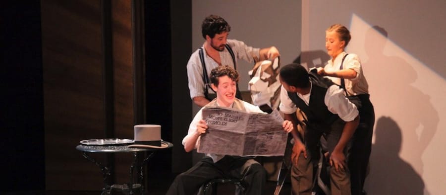 A group of four performers on stage interacting, with one person seated in a chair reading a newspaper in a theatrical setting.
