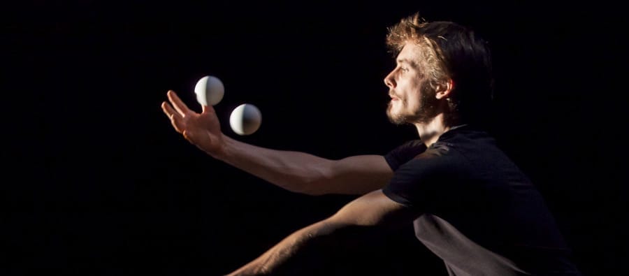 A juggler with brown hair, dressed in black t-shirt, skilfully balances white balls in motion against a dark background.