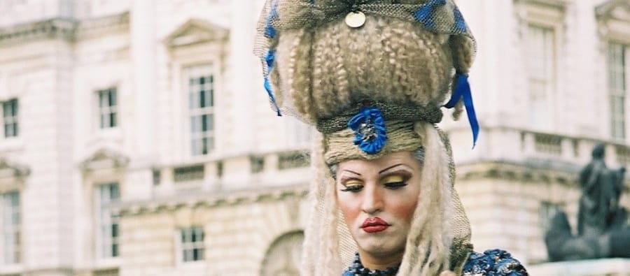 A performer in an extravagant blue and gold baroque-style costume styles a seated participant’s hair in an outdoor setting, with a historic building and a crowd in the background.