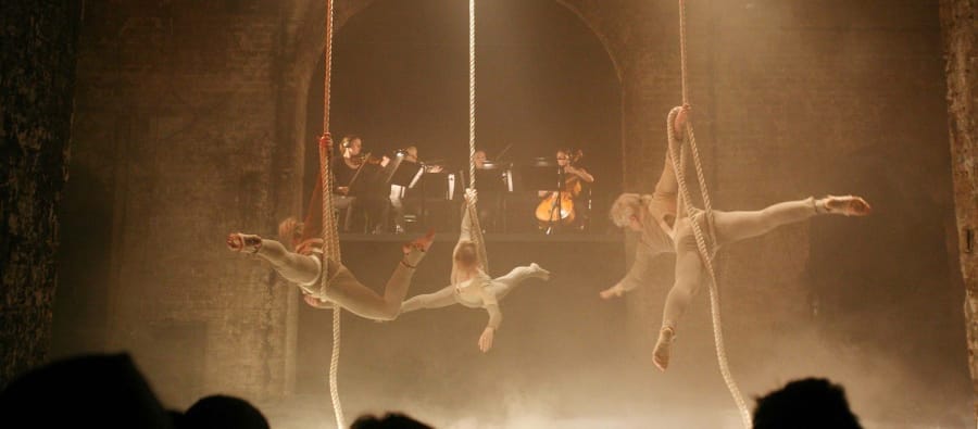 Three acrobats perform gracefully on ropes, as an orchestra plays in the background amid dramatic lighting and fog. Strings On Strings