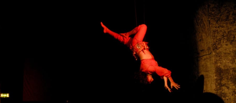 A performer in a red costume hangs upside down, gracefully suspended by strings against a dark backdrop. Layla Rosa What If