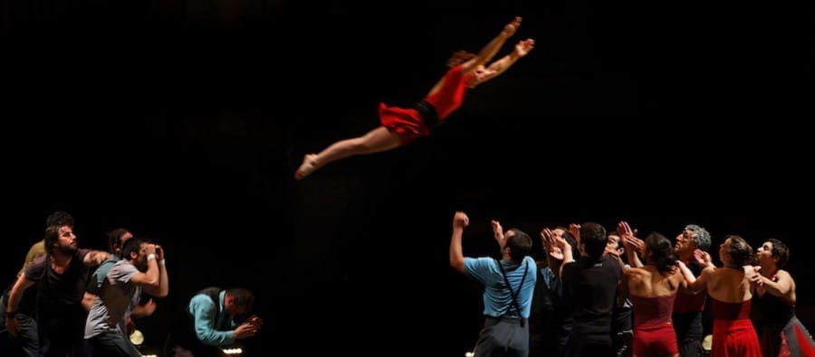 A dancer in a red dress flies gracefully through the air, leaping from one group of performers to another who lift them below, set in a dimly lit theatre. le grand C Company XY