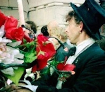 A man dressed in a black suit, white shirt, and a black top hat stands outside a building, surrounded by a large bouquet of red and white flowers, engaging with a crowd in the background.