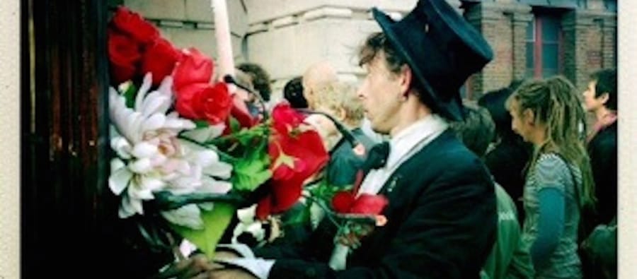 A man dressed in a black suit, white shirt, and a black top hat stands outside a building, surrounded by a large bouquet of red and white flowers, engaging with a crowd in the background.