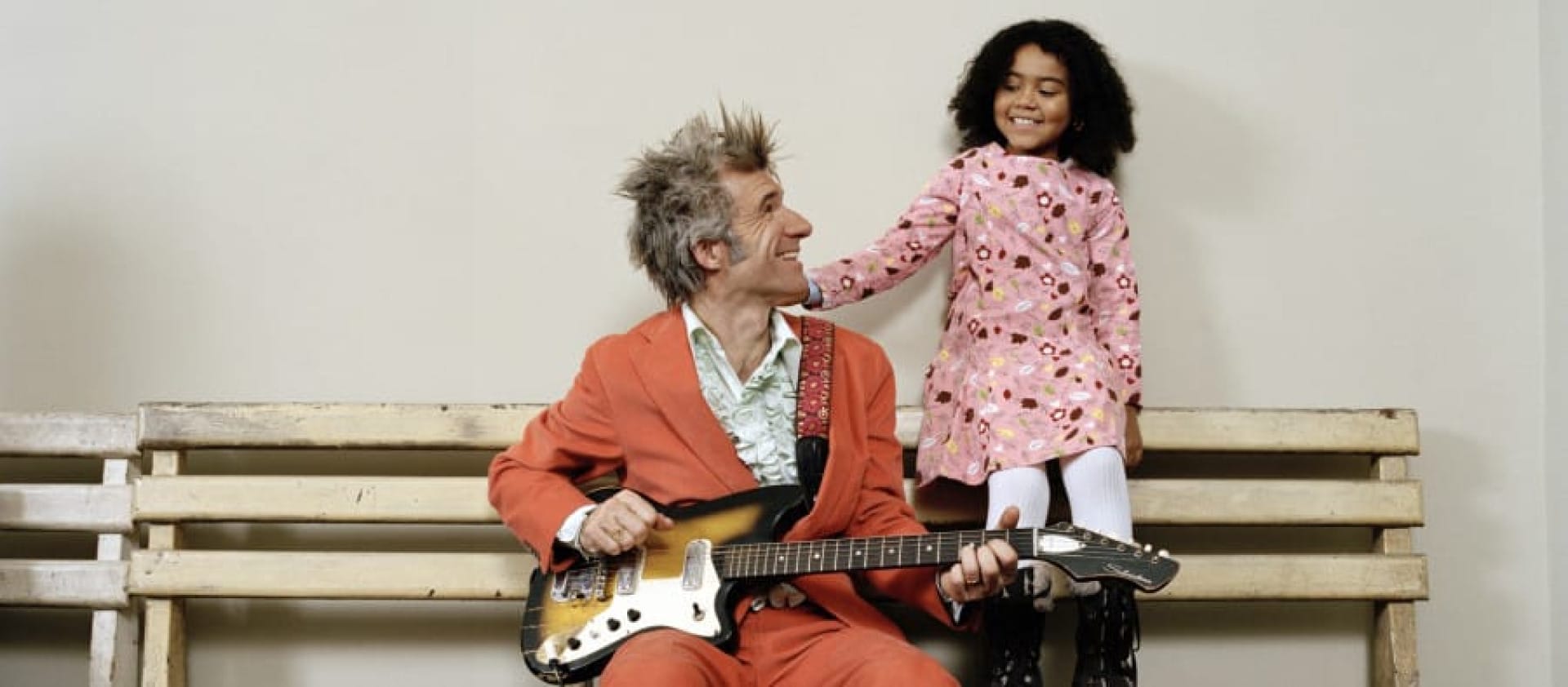 A musician in an orange suit sits on a bench, playing a guitar, while a child in a pink dress stands beside them, both smiling and looking at each other. Dan Zanes and Friends