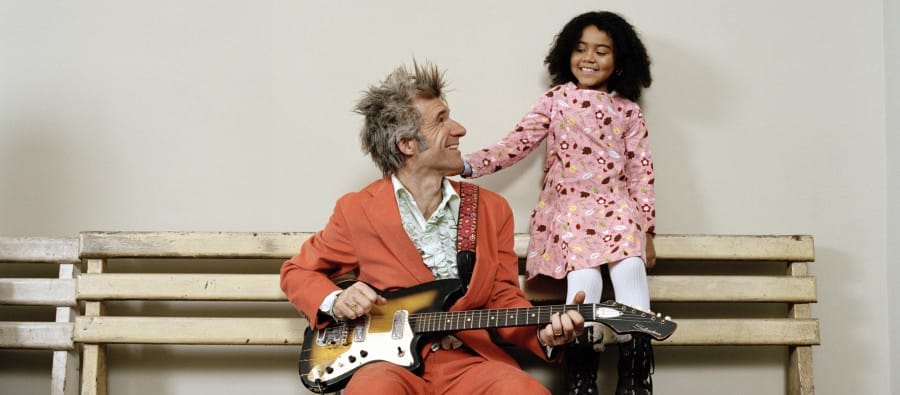 A musician in an orange suit sits on a bench, playing a guitar, while a child in a pink dress stands beside them, both smiling and looking at each other. Dan Zanes and Friends