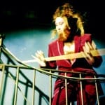 A performer in a deep red velvet suit with wild hair and a beard leans over a metal railing, holding a wooden plank, against a dimly lit blue background.