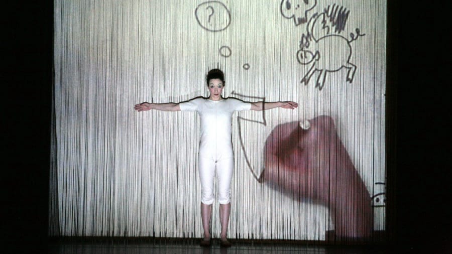 A performer dressed in a white bodysuit stands against a curtain-like screen, arms outstretched, as hand-drawn animated illustrations, including a flying pig and a question mark, are projected onto the backdrop. The image creates a playful and imaginative visual effect, blending live performance with animation.