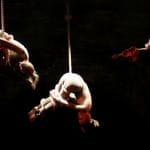 Three performers suspended from ropes, twisting and contorting, illuminated by dramatic lighting against a dark backdrop.