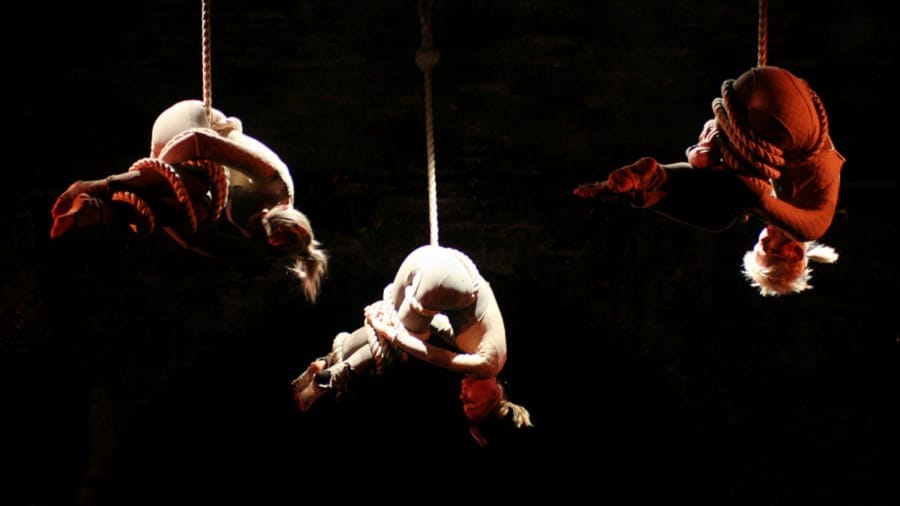 Three performers suspended from ropes, twisting and contorting, illuminated by dramatic lighting against a dark backdrop.
