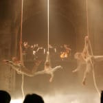 Three acrobats perform gracefully on ropes, as an orchestra plays in the background amid dramatic lighting and fog.