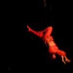 A performer in a red costume hangs upside down, gracefully suspended by strings against a dark backdrop.