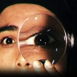 A close-up of a performer with dark hair and eyes on a black stage, peering through a circular magnifying glass held up to their eye What If…? | Layla Rosa © Ludo les Cognets, Max Ringham