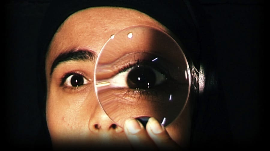 A close-up of a performer with dark hair and eyes on a black stage, peering through a circular magnifying glass held up to their eye What If…? | Layla Rosa © Ludo les Cognets, Max Ringham