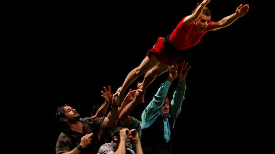 A performer in a red costume is gracefully airborne, supported by several individuals below in a dramatic, energetic pose against a dark backdrop.