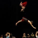 Two acrobats perform a high-flying aerial stunt above a group of performers who support them below, showcasing athleticism and teamwork in a dramatic theatre setting.
