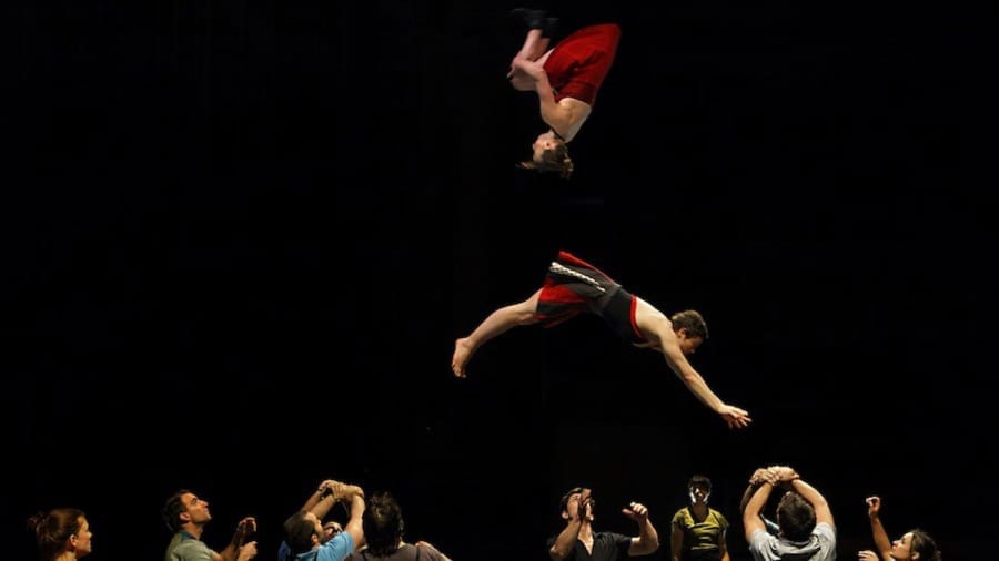 Two acrobats perform a high-flying aerial stunt above a group of performers who support them below, showcasing athleticism and teamwork in a dramatic theatre setting.