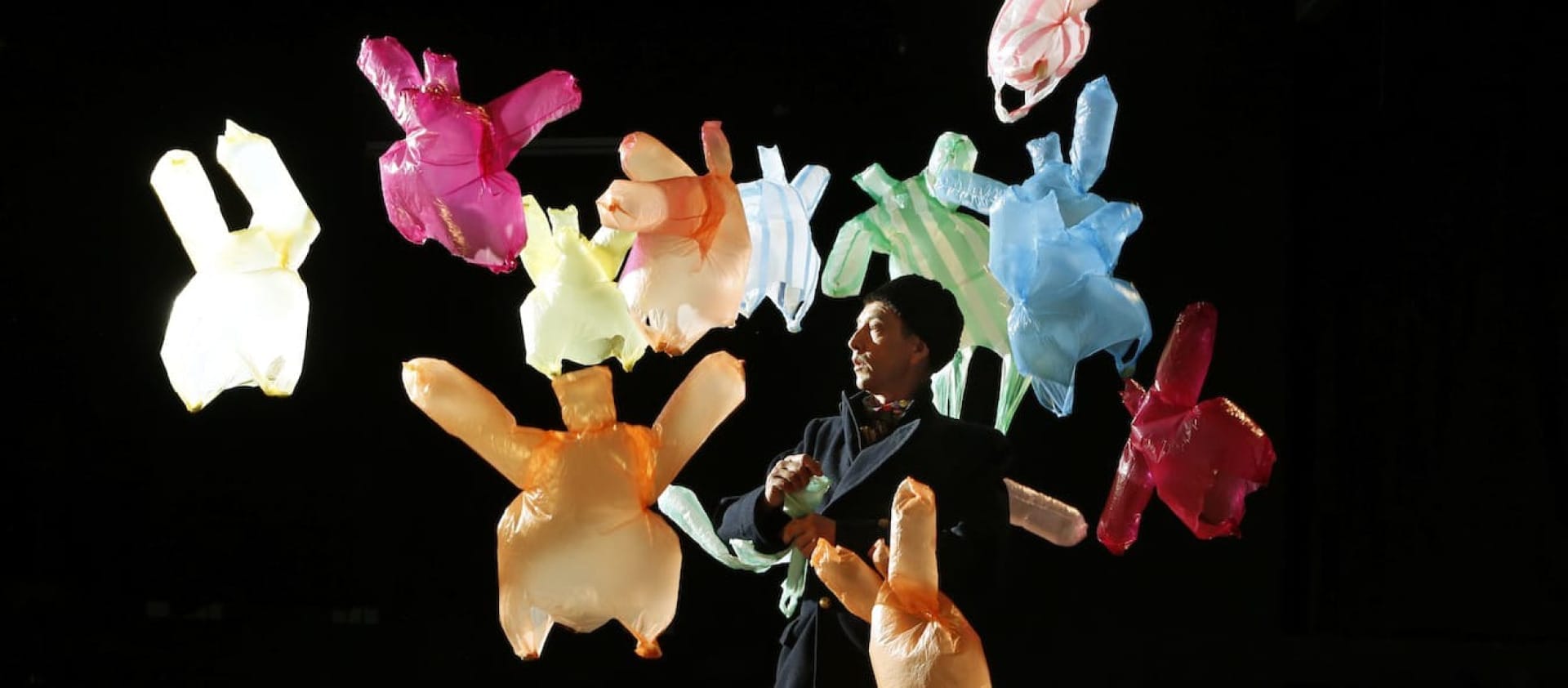 Many coloured plastic bags on a dark stage are being blown around by a floor fan, it looks as though they are dancing, one performer wearing black clothes and a hat stands in amongst the bags watching the action Our mission is to