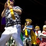 A lively performance on stage featuring a group of children and an adult with ukuleles, dressed in colourful outfits, enjoying the music.