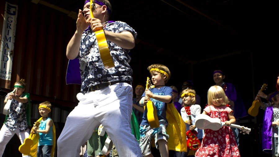 A lively performance on stage featuring a group of children and an adult with ukuleles, dressed in colourful outfits, enjoying the music.