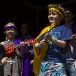 A group of children wearing colourful outfits and accessories play small musical instruments on stage, showcasing a joyful performance