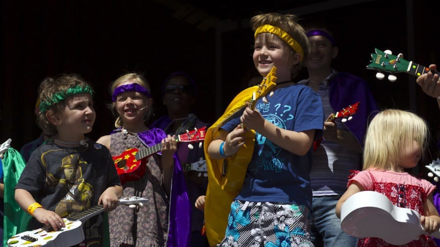 A group of children wearing colourful outfits and accessories play small musical instruments on stage, showcasing a joyful performance