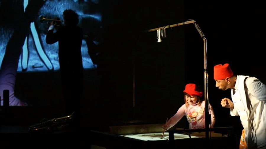 A performer in a white outfit and red fez interacts with a child in a pink shirt and red hat at a live performance, with a musician playing trumpet against blue screen in the background.