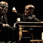 Two figures in black suits, with large bronze masks on their faces, sit at a wooden table, one raising a glass of beer, surrounded by a dark theatrical setting.