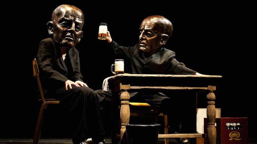 Two figures in black suits, with large bronze masks on their faces, sit at a wooden table, one raising a glass of beer, surrounded by a dark theatrical setting.