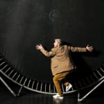 A person in casual attire leans against a large, round trampoline, surrounded by a dark stage backdrop.
