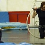A performer sits in a hoop suspended from the ceiling, while another person observes from the floor in a rehearsal setting.
