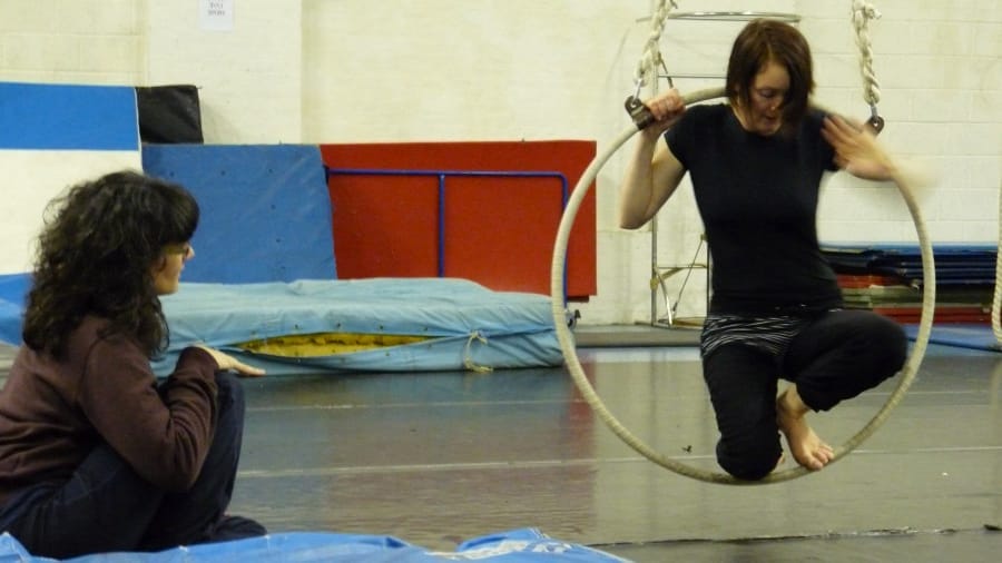 A performer sits in a hoop suspended from the ceiling, while another person observes from the floor in a rehearsal setting.