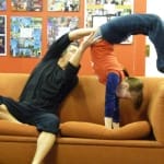 A performer balances another performer upside down in a playful acrobatic pose over an orange couch, with colourful posters seen in the background.