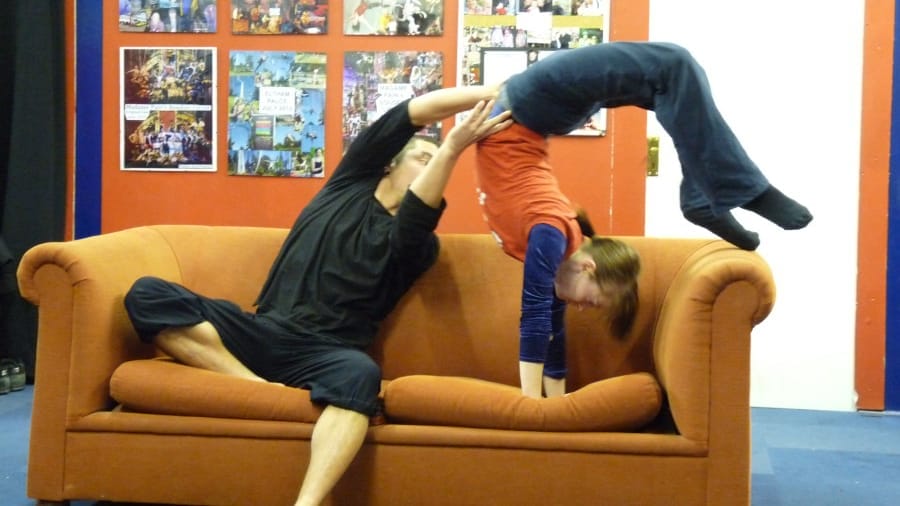 A performer balances another performer upside down in a playful acrobatic pose over an orange couch, with colourful posters seen in the background.