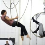 Two acrobats perform on aerial hoops, swinging elegantly in a rehearsal space.