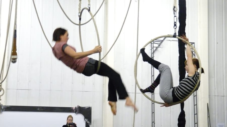 Two acrobats perform on aerial hoops, swinging elegantly in a rehearsal space.