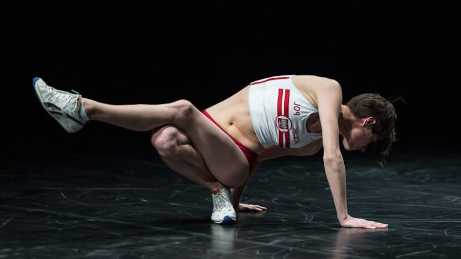 A dancer performs a dynamic pose on stage, showcasing strength and agility in athletic wear against a dark backdrop.