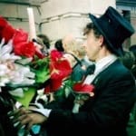 A man dressed in a black suit, white shirt, and a black top hat stands outside a building, surrounded by a large bouquet of red and white flowers, engaging with a crowd in the background.