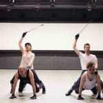Four performers in a dance studio strike dynamic poses, two riding on the others and wielding sticks, against a wide, blank screen backdrop.