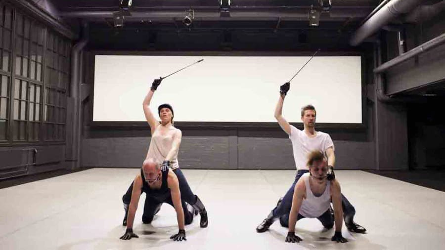 Four performers in a dance studio strike dynamic poses, two riding on the others and wielding sticks, against a wide, blank screen backdrop.