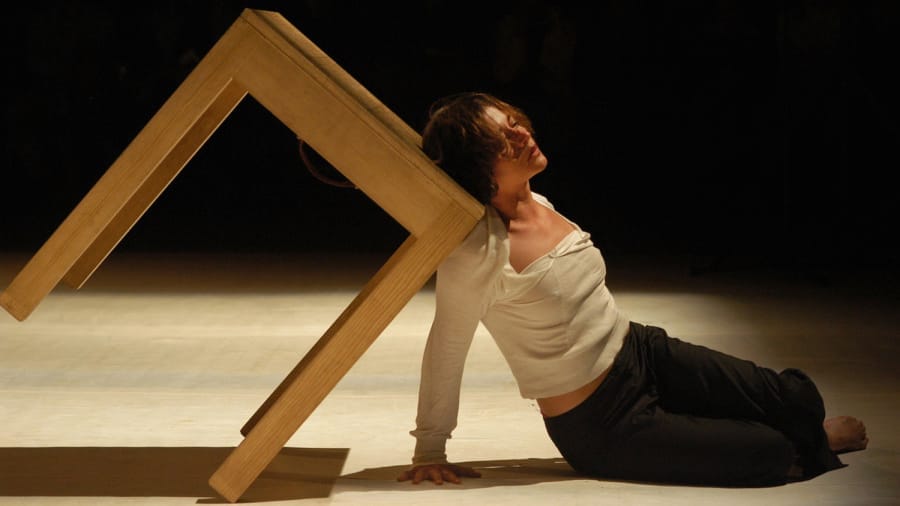 A dancer leans against a wooden table, suspended diagonally on two legs, creating an intimate and dynamic pose on a dimly lit stage.