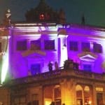 Illuminated historic building at night, featuring purple lights and people on the roof, creating a festive atmosphere.