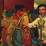 A man wearing a paint-splattered white coat and a beret stands next to a colourful cutout of a king holding a sceptre, blending himself into the artwork in a playful interaction.