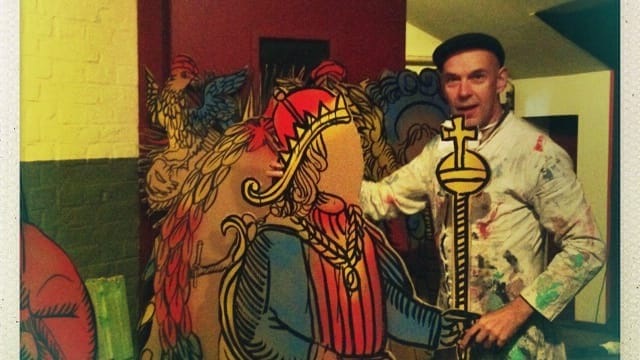 A man wearing a paint-splattered white coat and a beret stands next to a colourful cutout of a king holding a sceptre, blending himself into the artwork in a playful interaction.