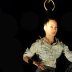 A close-up of a performer suspended by their hair from a metal ring, gazing calmly at the camera against a dark backdrop.