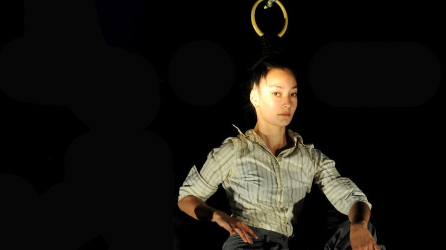 A close-up of a performer suspended by their hair from a metal ring, gazing calmly at the camera against a dark backdrop.
